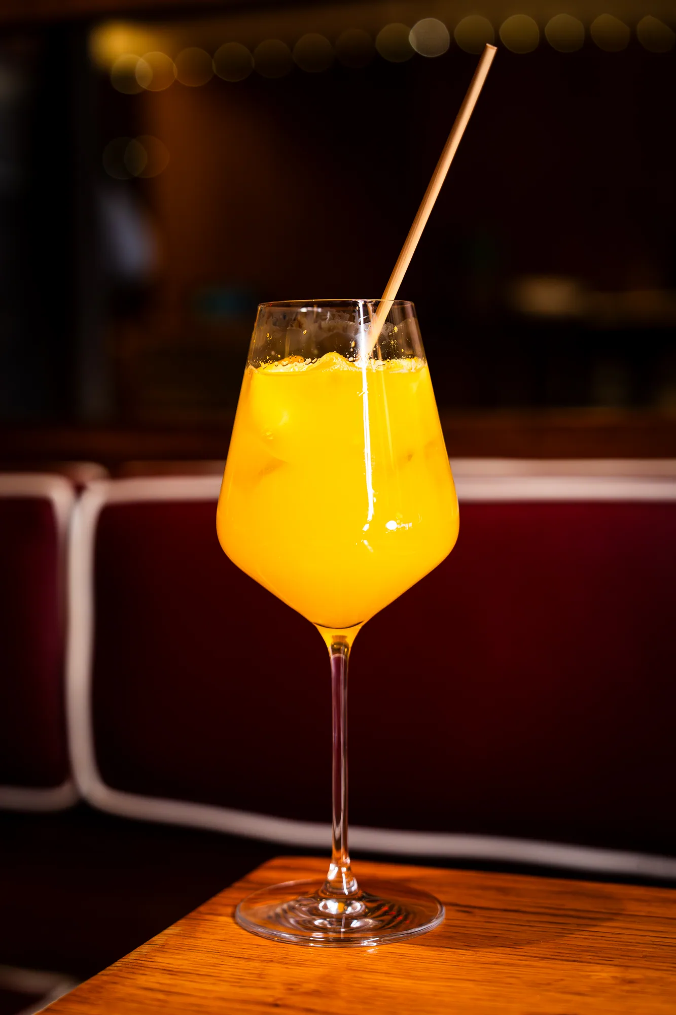 An orange spritz in a wine glass on the wooden table beside a red banquette