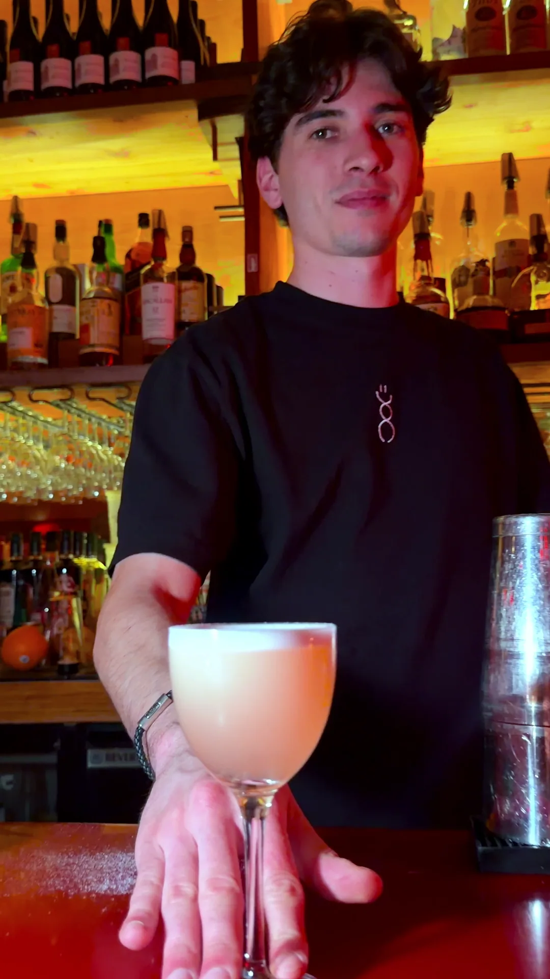 A bartender holding out a coupe of pale cocktail over the red bar top, bottles glowing amber behind