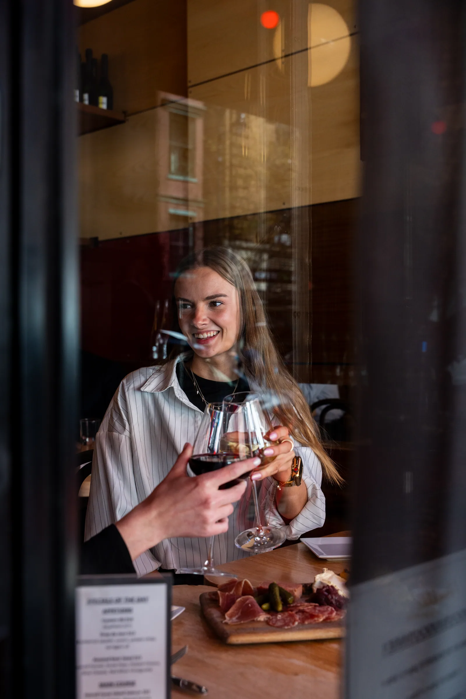 A guest with a glass of red wine and a charcuterie board, seen through the front window of Amélie West Village